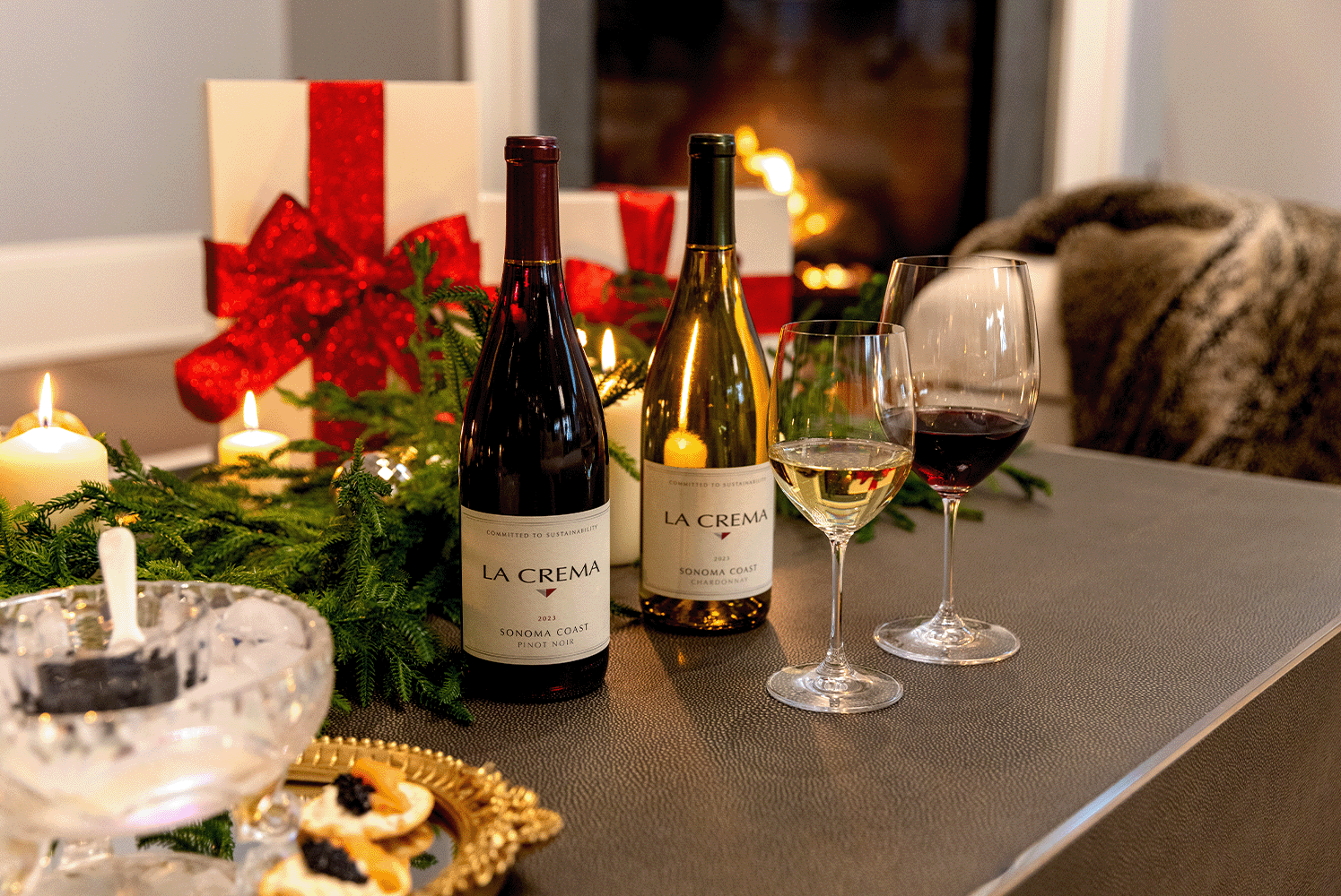 Holiday table with La Crema chardonnay and pinot noir bottles and filled glasses of wine with gift boxes and roaring fireplace in the background