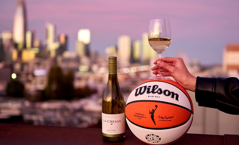 A glass of La Crema Chardonnay, bottle of wine, and a Wilson WNBA basketball with a sunset over a city skyline